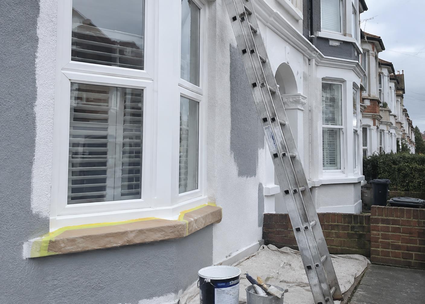 Exterior painting in progress on a rendered terraced house with a ladder and masked window sill